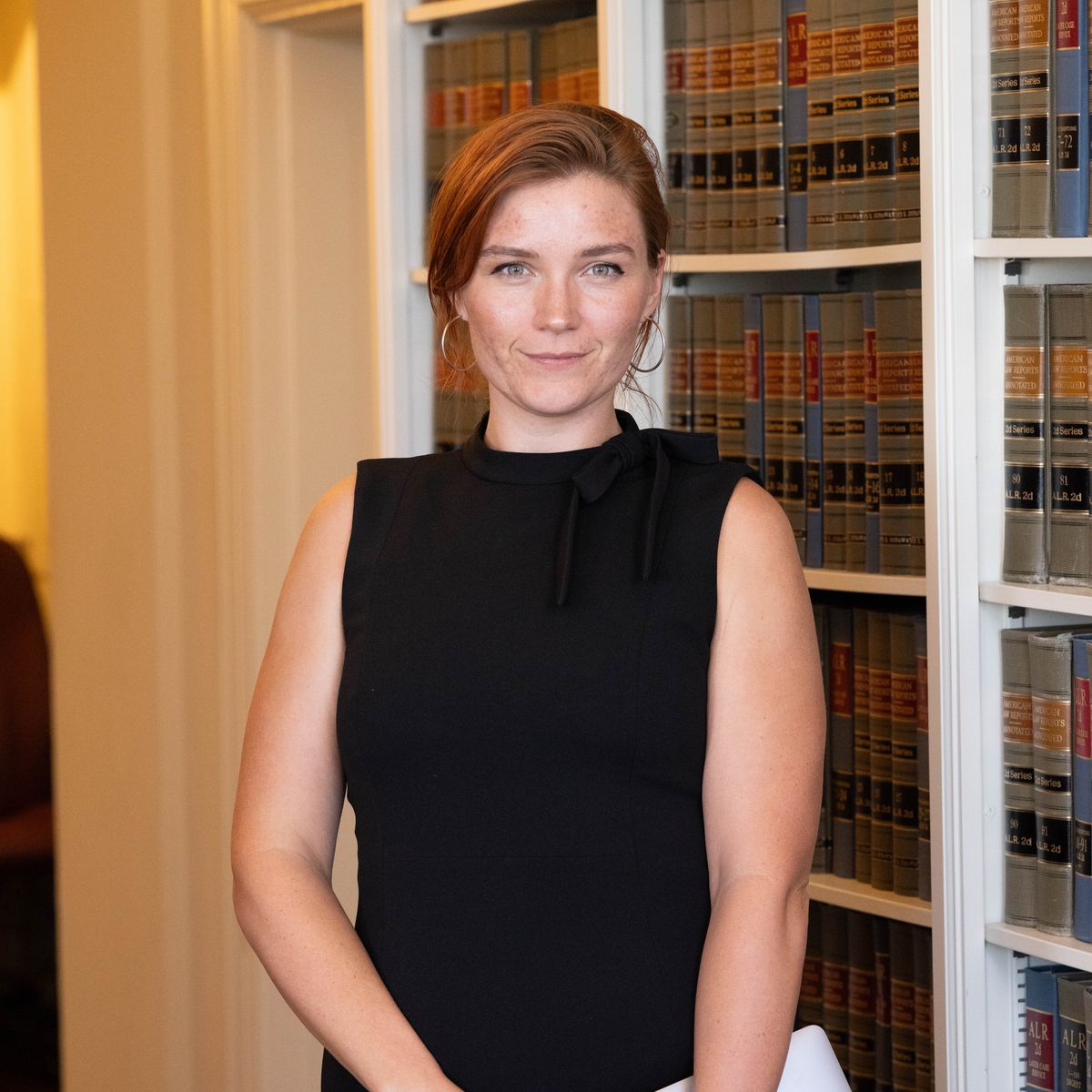 red-headed woman standing in front of a bookshelf looking serious